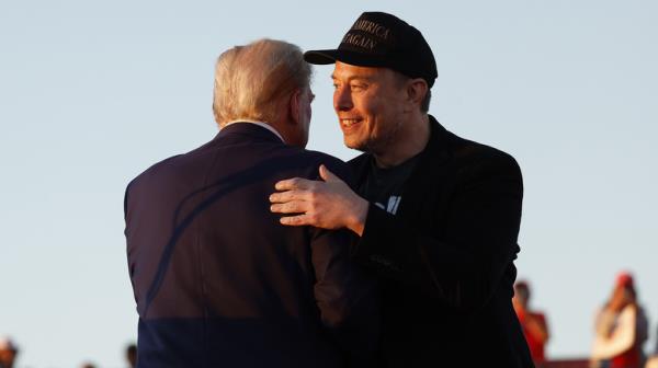 Elon Musk in dark ba<em></em>seball cap hugging Do<em></em>nald Trump on stage