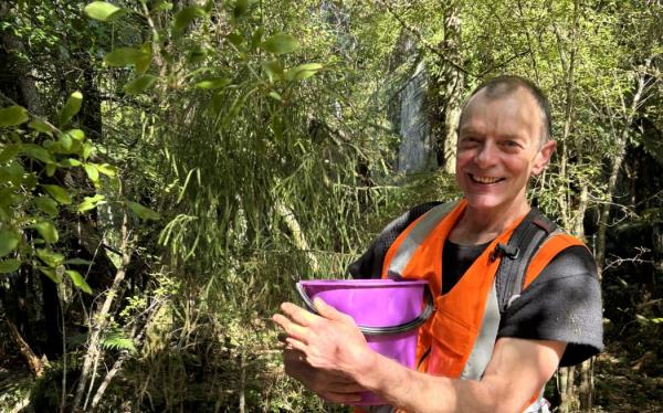 A man in a t-shirt and orange hi-vis vest holds a purple bucket and smiles at the camera, standing in lush green forest.