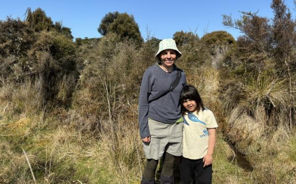 A woman in a bucket hat, long-sleeved top and waders stands next to a small child with dark hair and a yellow t-shirt. They are standing in front of brown scrubby wetland vegetation beneath a blue sky, and are smiling at the camera.