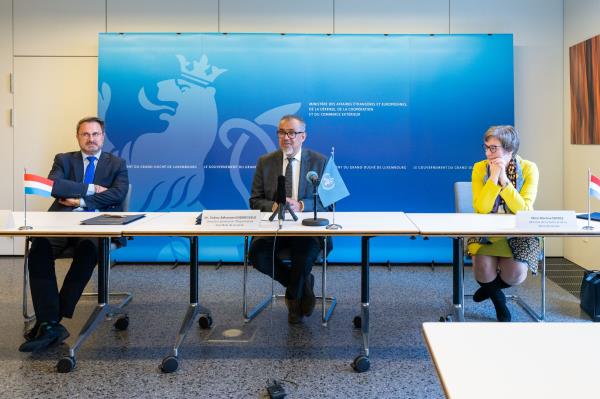 Two men and a woman sitting at a large table in a room, with small sized flags of Luxembourg and the Worls Health Organization standing on the table.